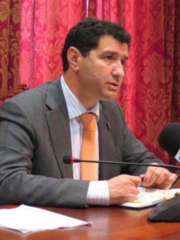 Un hombre en traje hablando en una conferencia de prensa, con un fondo de cortinas rojas y un micrófono en la mesa. La imagen parece ser de un evento formal o político.