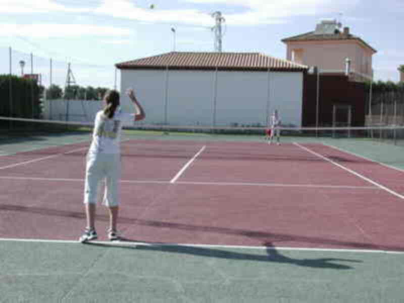 Dos personas juegan tenis en una cancha de tenis con un edificio en el fondo. La imagen muestra a una persona preparándose para golpear la pelota con un raqueta en una cancha de tenis.