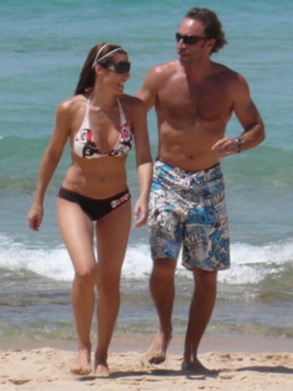 Un hombre y una mujer caminan en la playa, con el mar en el fondo. La mujer lleva un bikini y el hombre usa pantalones cortos de playa con estampado.