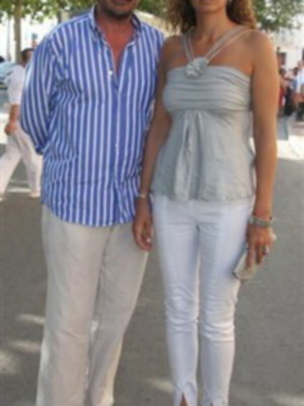 Una pareja camina por una calle con ropa casual. La mujer lleva un top gris y pantalones blancos, mientras que el hombre usa una camisa a rayas azul y blanco con pantalones blancos. Ambos llevan sandalias de color claro.