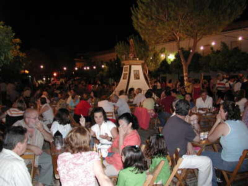 Una feria nocturna con gente sentada en mesas bajo un cielo oscuro. La escena está iluminada por faroles y luces de la feria, creando una atmósfera festiva. La gente está disfrutando de la noche, con algunos conversando y otros observando el ambiente.