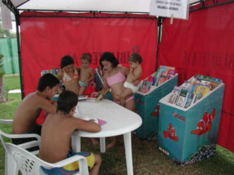 Una familia disfruta de un día en la playa, con niños leyendo libros bajo una sombrilla roja.