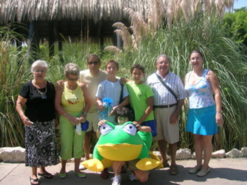 Una familia de siete personas está posando frente a una figura inflable de un sapo verde y amarillo. La imagen se encuentra en un entorno natural con vegetación alta y una estructura de techo de paja al fondo.