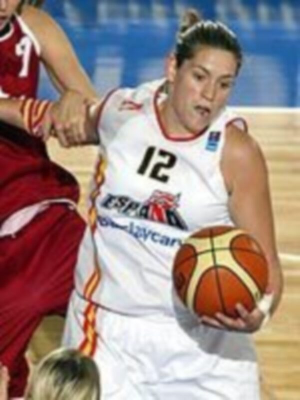 Una jugadora de baloncesto con el número 12 en la camiseta blanca sostiene un balón de color naranja y rojo. Está en una cancha de baloncesto, con otra jugadora de la selección española detrás.