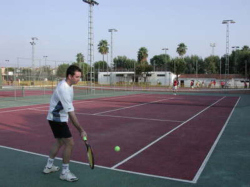 Un hombre juega tenis en una cancha de tenis con un palo de tenis en la mano y una pelota en el aire. El fondo muestra un paisaje con árboles y una estructura de luz en el horizonte.