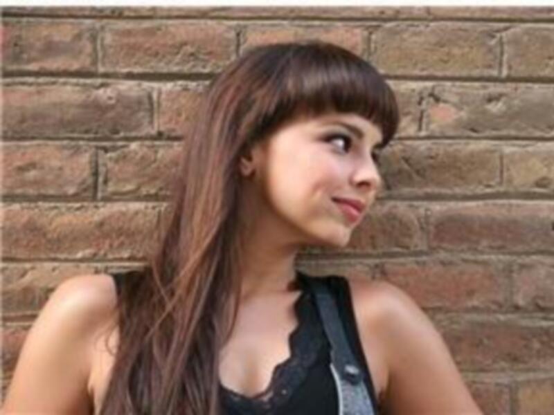 Una mujer con cabello largo y ondulado, mirando hacia la derecha. Se encuentra frente a una pared de ladrillos.