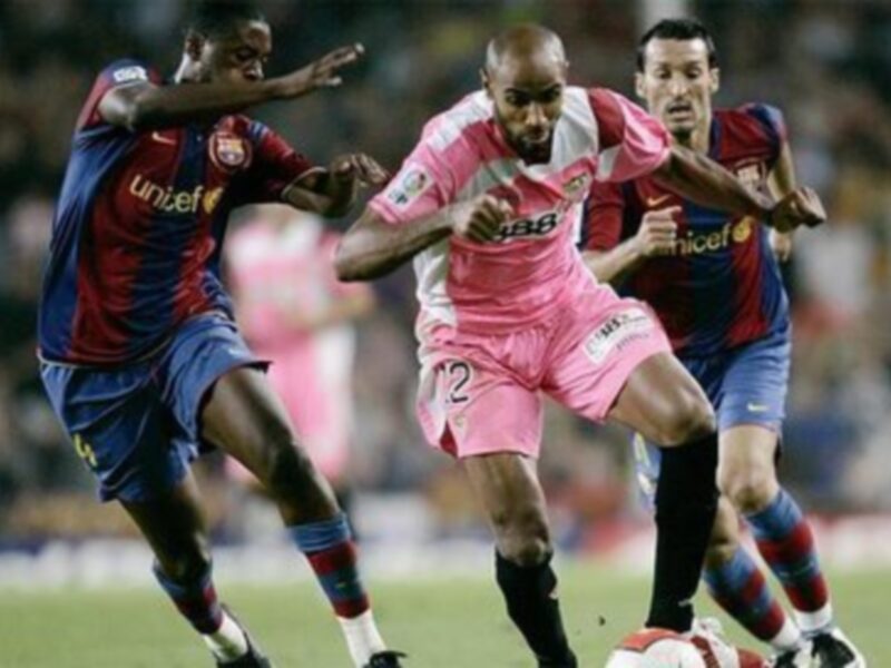 Imagen de un partido de fútbol donde dos jugadores del Barcelona, uno con camiseta roja y otro con camiseta azul, intentan controlar el balón. El jugador en rosa es el centro de atención, corriendo y luchando por el control del balón.