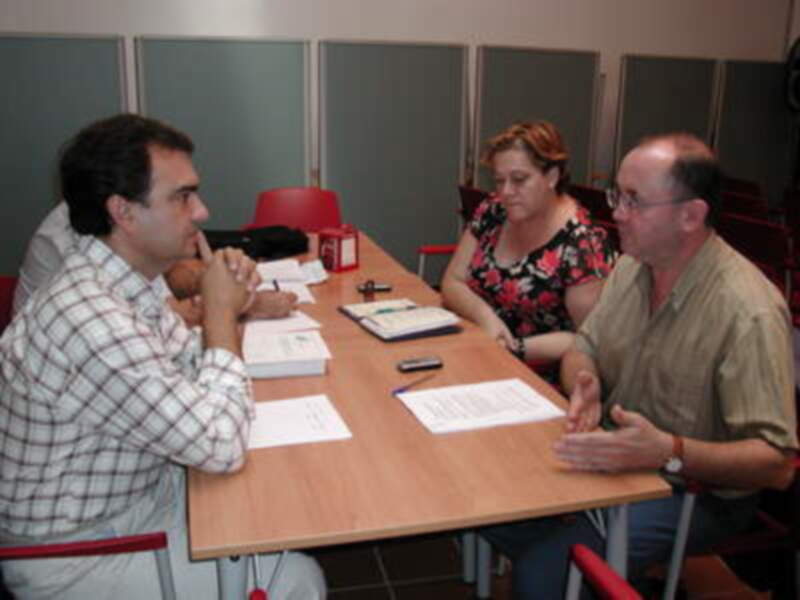 Tres personas sentadas en una mesa con documentos y un teléfono móvil. La imagen parece ser de una reunión o conferencia.