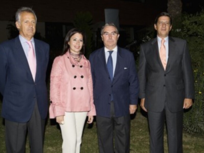 Cuatro personas posan juntas en un ambiente formal, tres de ellos vestidos con trajes y sombreros, mientras que la cuarta lleva una chaqueta rosa. La escena parece ser nocturna, con luces de fondo y vegetación alrededor.