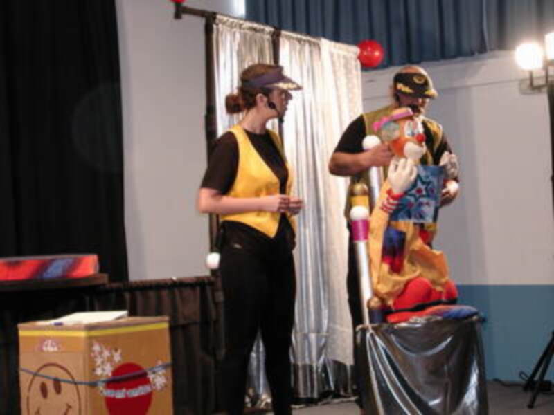 Dos personas en un escenario, una con un disfraz de payaso y la otra con un traje negro y amarillo. Hay una figura de juguete en el centro, con un globo rojo y una pelota blanca. La escena está iluminada con luces de teatro.
