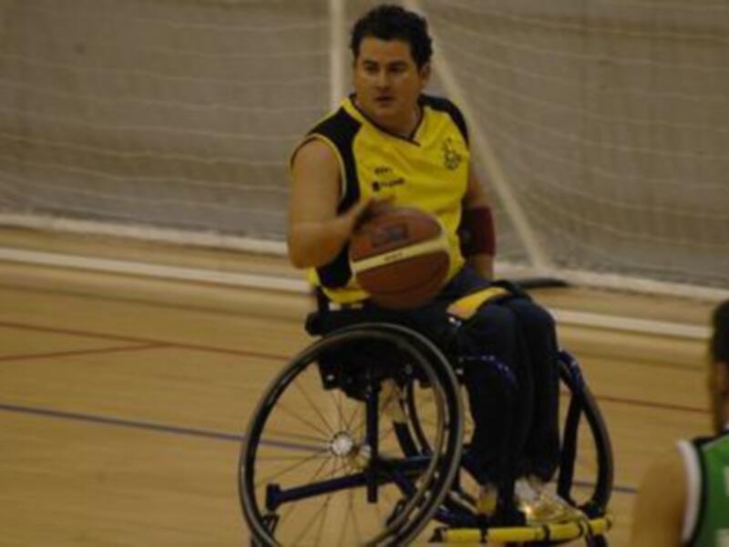 Un hombre en silla de ruedas juega baloncesto. El fondo es una cancha deportiva con un objetivo de portería visible.