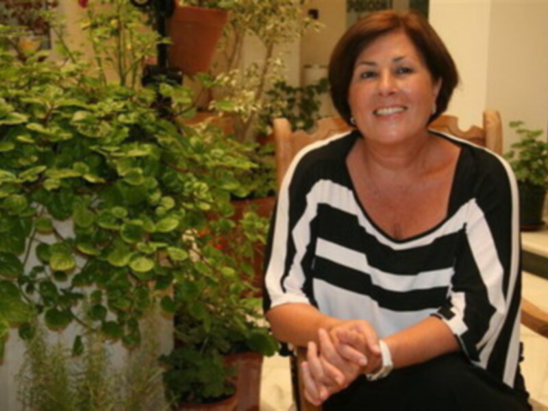 Una mujer sonríe mientras se sienta en una silla de madera frente a un escritorio. A su lado, hay plantas verdes y flores en macetas. La imagen transmite una sensación de calma y bienestar.