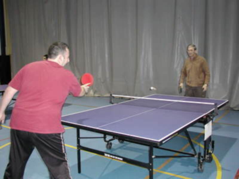 Un hombre juega a ping pong frente a una cortina oscura. El tablero de ping pong es azul con líneas blancas y amarillas en el suelo. El hombre está vestido con una camiseta roja y pantalones de chándal negros.