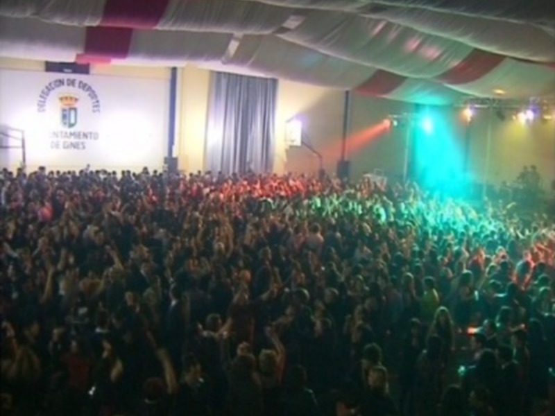 Una multitud de personas en un auditorio, con luces de color verde y rojo iluminando la escena. El fondo muestra un cartel con texto en español, probablemente el nombre del evento o organización.