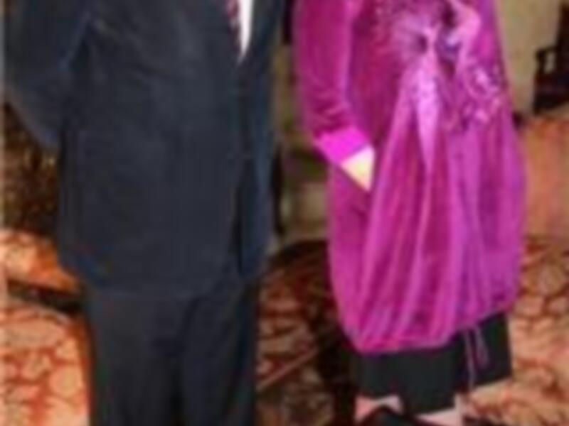 El hombre lleva un traje negro con corbata roja y la mujer una chaqueta de terciopelo morado. Ambos están en un entorno formal con una alfombra y un espejo visible en el fondo.