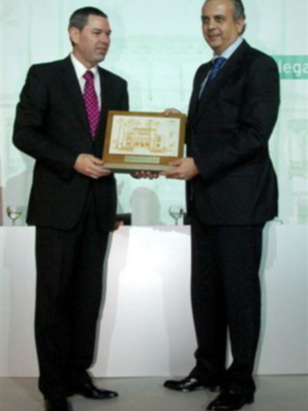 Dos hombres en trajes sostienen un certificado. El fondo es una pantalla con texto y imágenes gráficas.