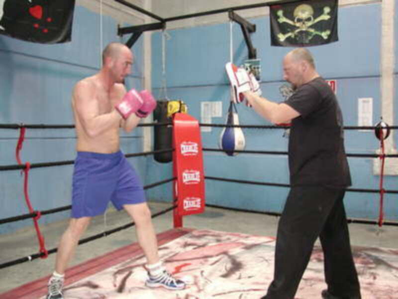 Un boxeador entrenando con un entrenador en una sala de boxeo, con un fondo azul y una bandera con un cráneo y un puño en la pared.