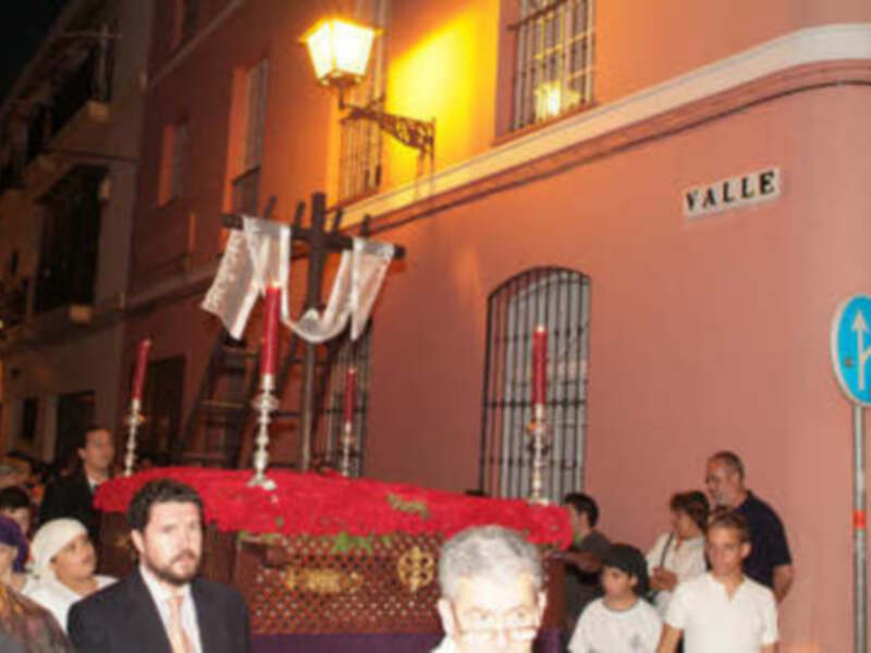 Una procesión religiosa en una calle de la ciudad, con personas vestidas formalmente y un carro adornado. La imagen muestra detalles como farolas, una calle con edificios de color rojo y un cartel que dice "Valle".