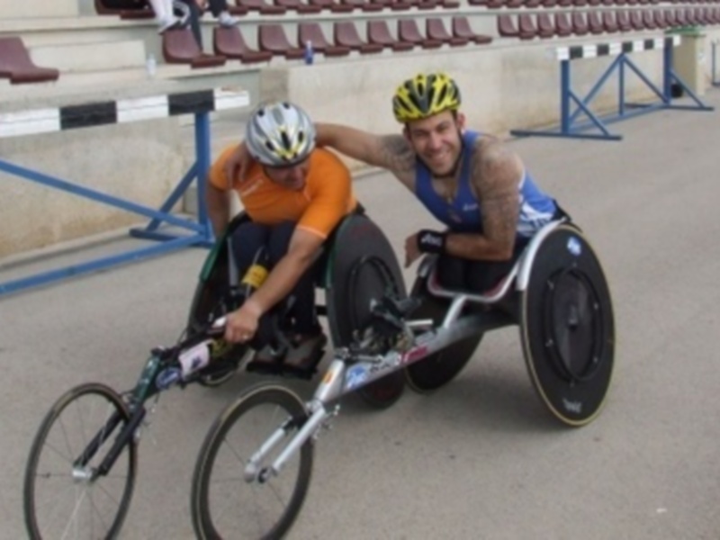 Dos atletas en sillas de ruedas compiten en un evento de atletismo adaptado. El ambiente es una pista deportiva con gradas vacías en el fondo.