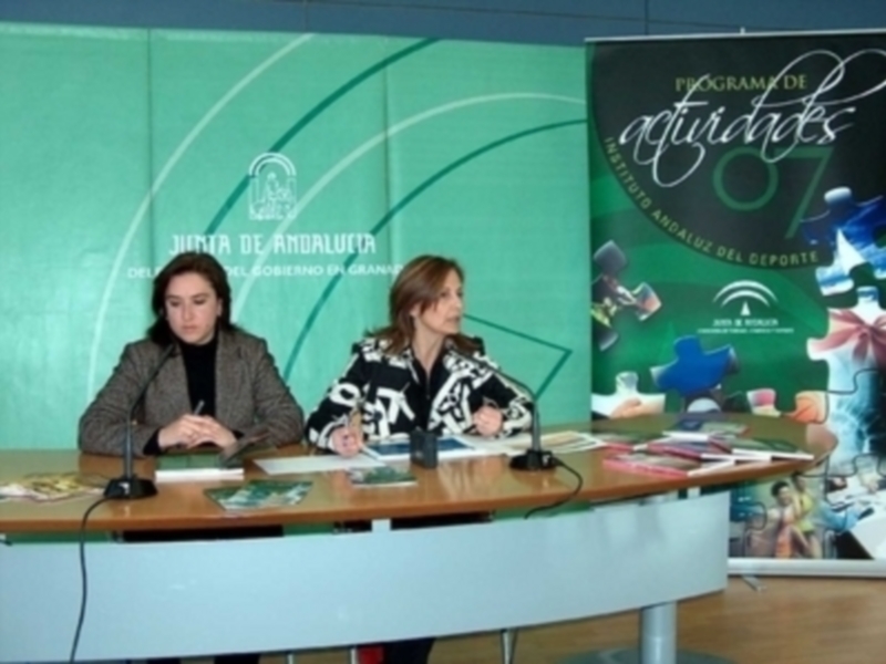 Dos mujeres sentadas en una mesa de conferencia, frente a un fondo verde con el logo del Junta de Andalucía y un cartel que anuncia actividades deportivas.