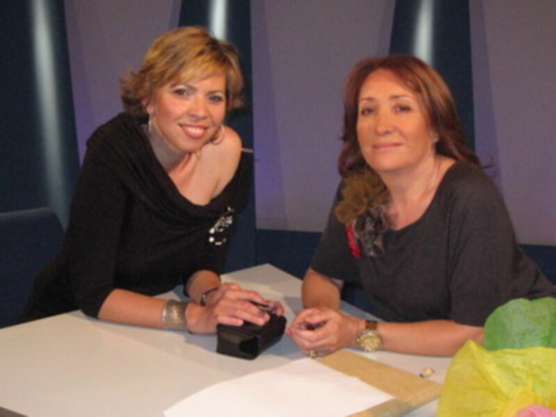 Dos personas sonrientes en un estudio, una con cabello corto y la otra con cabello largo. Ambas están sentadas en un escritorio, una sosteniendo un objeto negro. La imagen sugiere un entorno de trabajo o presentación.