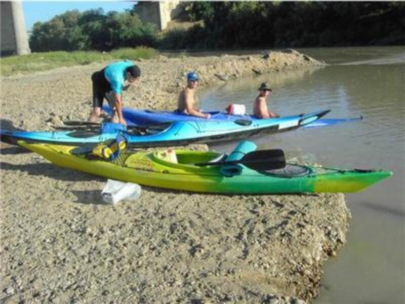 Tres personas preparándose para una actividad acuática en una orilla de río, con dos kayaks colgados en el suelo.