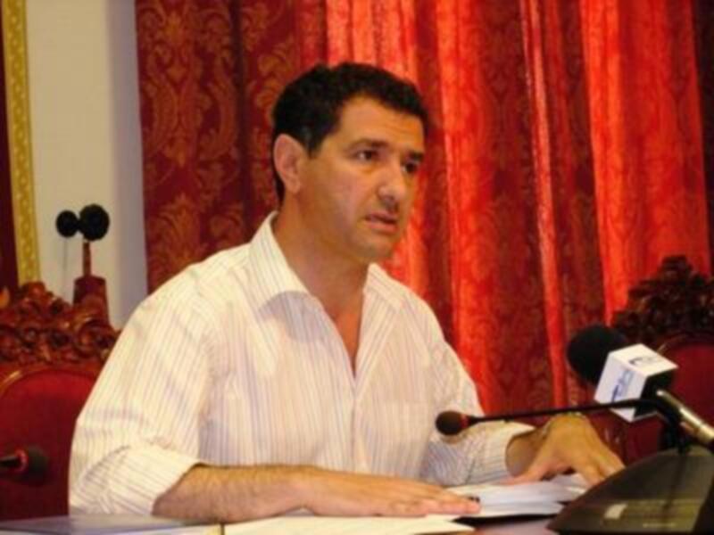Persona hablando en un evento formal con micrófono y documentos en la mesa.