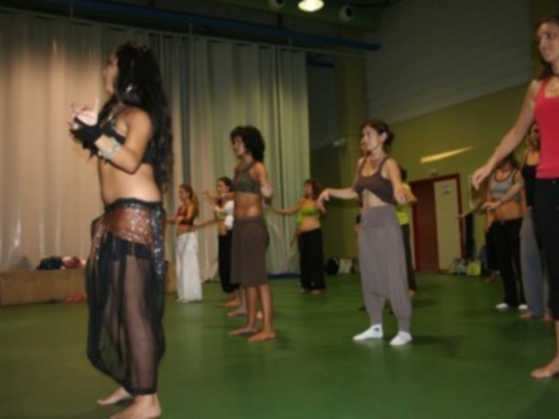 Una escena en un gimnasio con varias personas bailando. La imagen muestra a una mujer en el centro, vestida con un atuendo de baile, y otras personas alrededor. La iluminación es suave y la pared de fondo está decorada con cortinas.