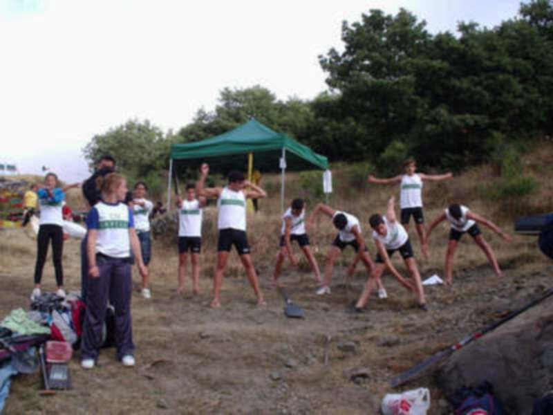 Una escena de una entrenamiento de atletismo en un campo de entrenamiento, con atletas realizando estiramientos y una carpa de protección en el fondo.