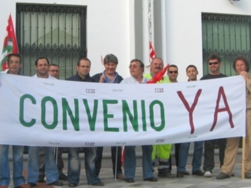 Un grupo de personas sostiene un cartel que dice "CONVENIO YA" frente a una estructura con barras verticales.