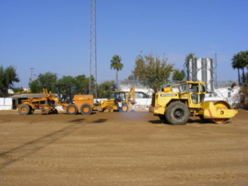 Tres vehículos de construcción trabajando en un campo de fútbol, con una torre y un tanque de agua al fondo.