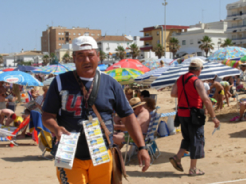 Un hombre con una camiseta azul y un sombrero blanco sostiene un cartel en la playa, rodeado de otros bañistas y sombrillas coloridas.