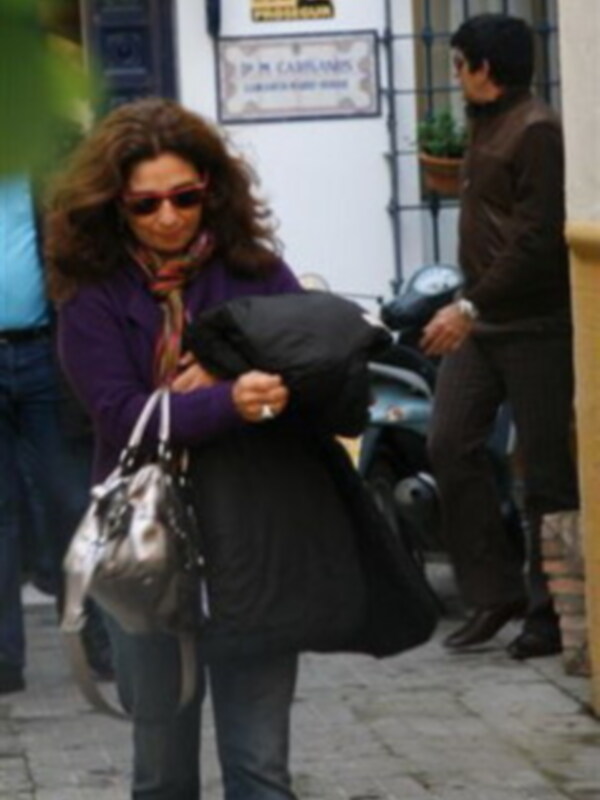 Una mujer con gafas de sol y una chaqueta púrpura camina por la calle, seguida por un hombre con una chaqueta marrón. Detrás de ellos hay una puerta con un cartel amarillo y negro, y un coche azul parado en el fondo.