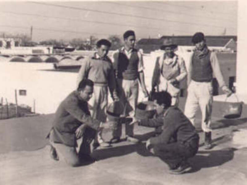 Grupo de personas en una terraza, posiblemente en un edificio antiguo. Algunos llevan maletas y otros están sentados, con un fondo de edificios y cielo despejado.