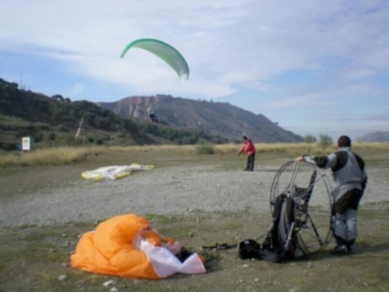 Un parapente verde volando sobre un paisaje de montaña, con una persona preparándose para el vuelo y otra con su parapente en tierra.