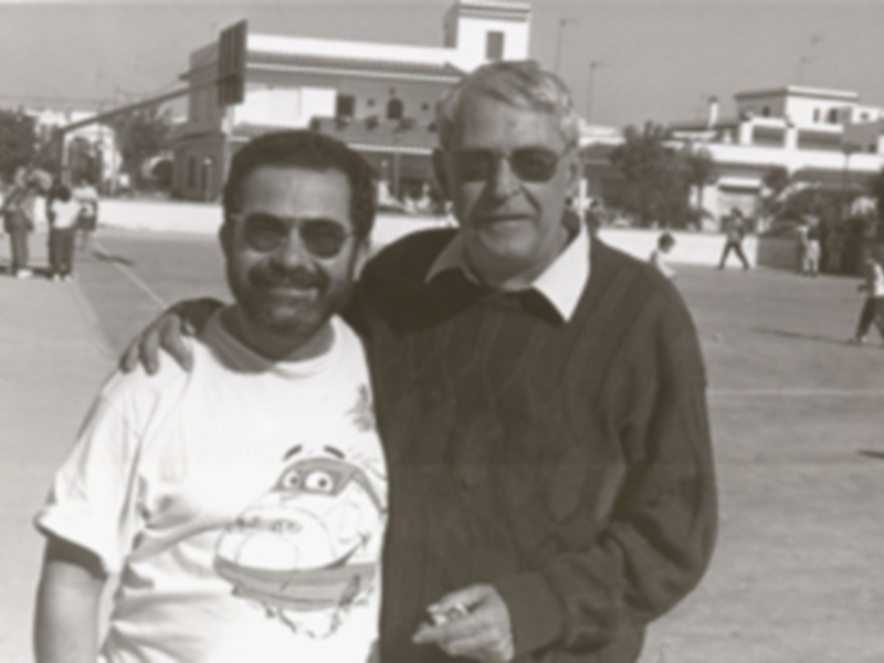 Dos hombres sonrientes en una calle, uno con camiseta de dibujos animados y el otro con jersey negro. Fondo urbano con edificios y personas en la distancia.