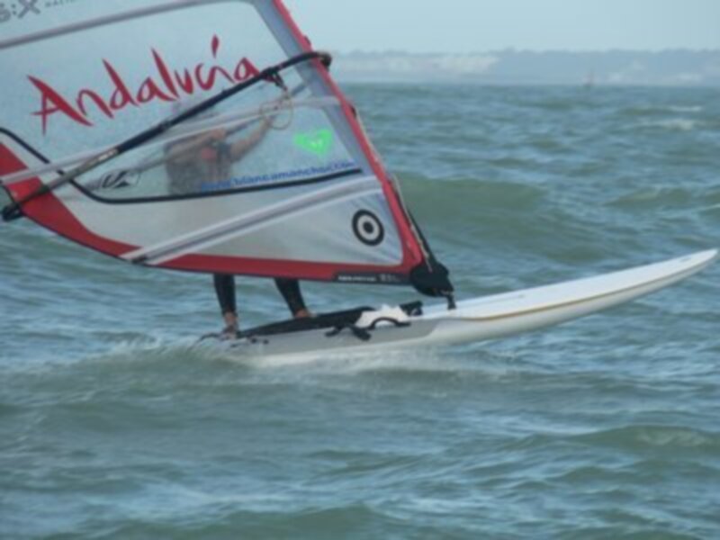 Un windsurfer navega sobre el agua con una vela que muestra la bandera de Andalucía. La imagen captura el movimiento y la emoción del deporte en un entorno natural con una costa visible al fondo.