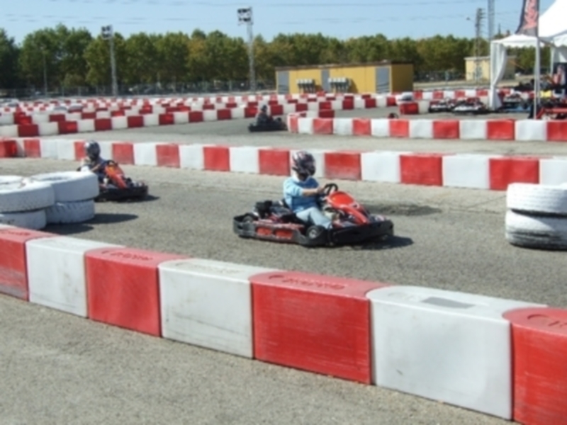 Una escena de karting con niños conduciendo go-karts en un circuito rodeado por barreras rojas y blancas. El cielo es claro y el paisaje está lleno de árboles verdes en la distancia.