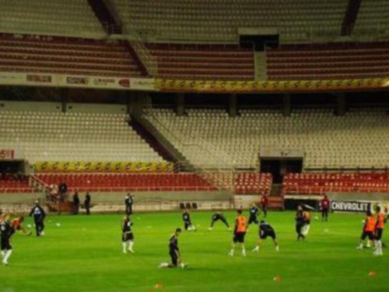 Un estadio vacío con asientos rojos y amarillos. Jóvenes entrenando en el campo de fútbol, con balones dispersos y varios espectadores observando.