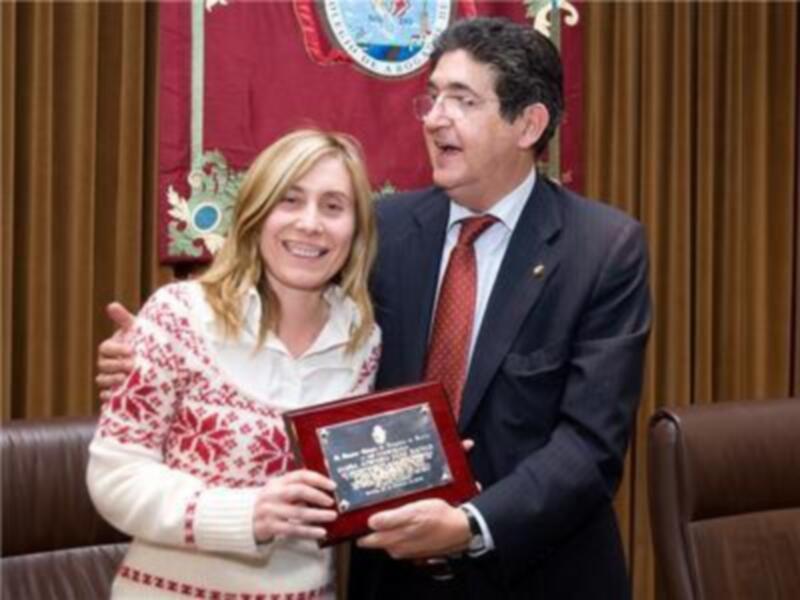 Una mujer sonríe mientras sostiene un diploma, junto a un hombre en traje que también sonríe. El fondo muestra una bandera y un escudo de armas, indicando un ambiente oficial o institucional.