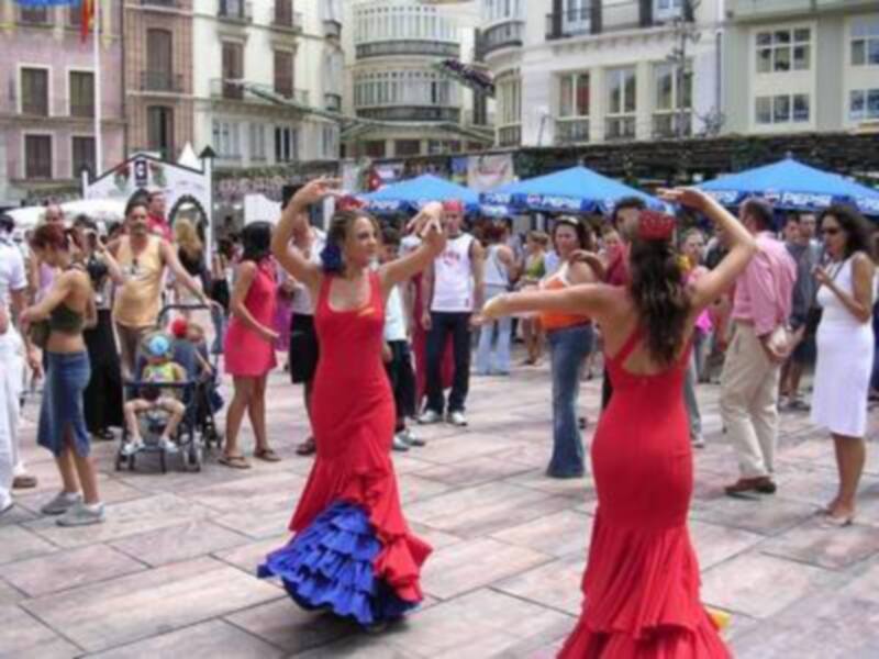 Damas bailando flamenco en una calle de la ciudad. Hombre con sombrero y niño en silla de ruedas a su lado. Fondo urbano con edificios y gente observando la danza.