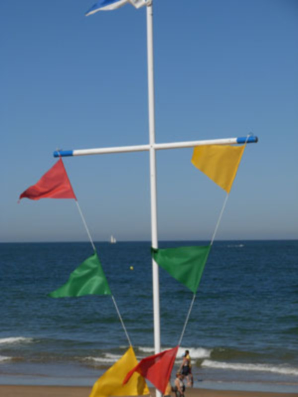 Banderas de colores ondean en un poste blanco frente a la playa, con el mar y una embarcación lejana al fondo.