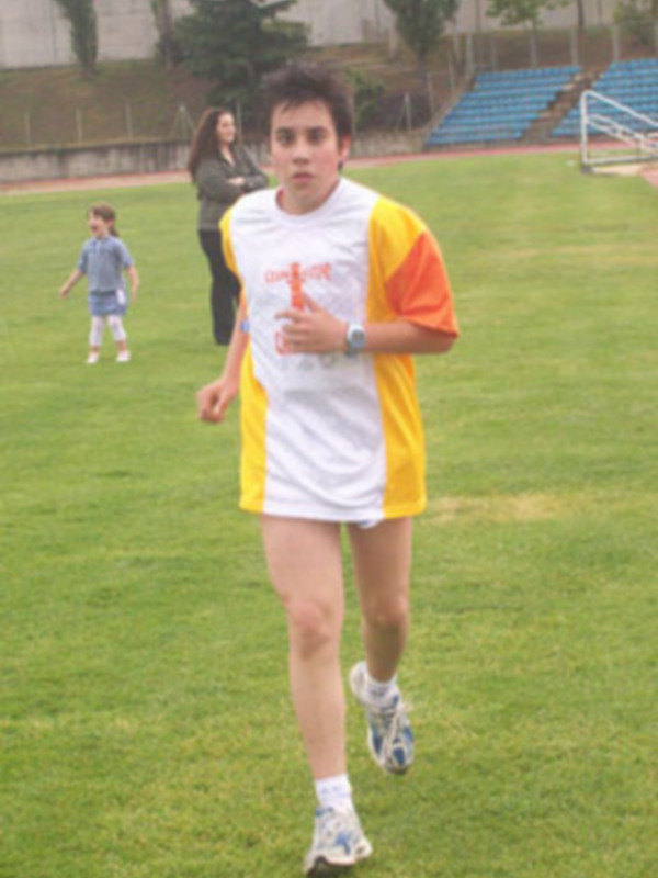 Un joven corriendo en un campo de césped, con una camiseta amarilla y blanca y pantalones cortos. Detrás de él, una niña en un uniforme azul y otro niño en el fondo. La escena está en un día soleado, con una persona observando desde atrás.