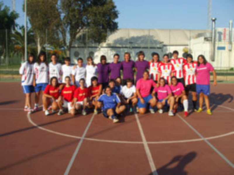 Equipo de fútbol femenino posando para una foto de equipo en un campo de fútbol al aire libre. El equipo está dividido en dos equipos, uno con camisetas rojas y otro con camisetas moradas y amarillas. El fondo muestra un edificio blanco y un césped verde.