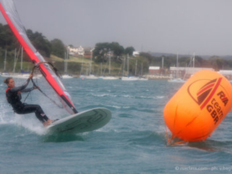 Un windsurfer navega sobre el agua azul, con una vela roja y negra en el aire. A su lado, un globo naranja con la marca "RA" y "LGBA" flota en el agua. En el fondo, se observan edificios y árboles alineados con una línea de costa.
