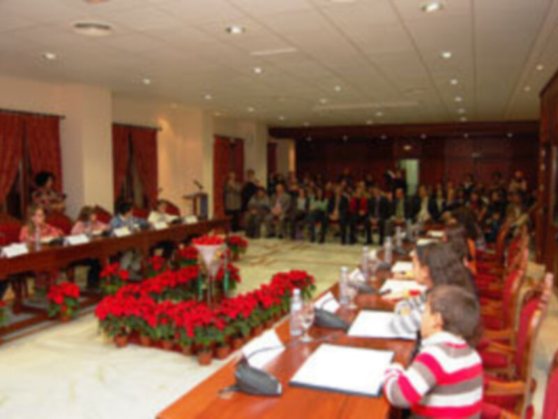 Salón con mesas llenas de flores rojas, gente sentada en filas y una pantalla en el fondo.