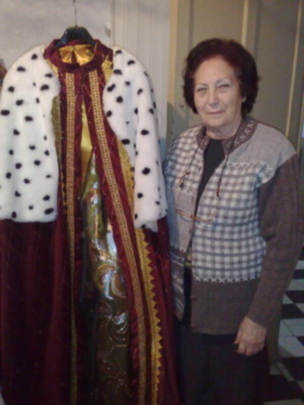 A una mujer posando junto a un traje histórico. El traje tiene un chaleco blanco con puntos negros y una capa roja con detalles dorados. La mujer está vestida con un suéter a cuadros y una falda negra. La escena parece ser interior, posiblemente en un museo o exposición histórica.