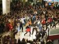 Una multitud de personas bailando en un centro comercial, con carteles y publicidad al fondo.