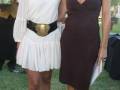 Dos mujeres posan en un evento formal, una con vestido blanco y cinturón dorado, la otra con vestido negro. El fondo muestra un ambiente verde y soleado.