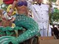 Una figura de una sirena con cola verde y escamas, junto a un hombre vestido como capitán, sosteniendo un tridente.